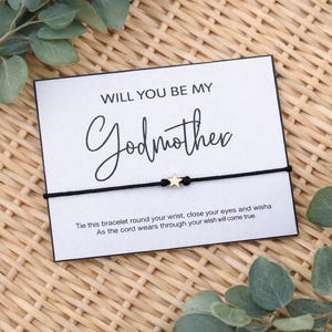 May include: A white card with the text "WILL YOU BE MY Godmother" and a black cord bracelet with a gold star charm. The card is on a woven surface with green leaves.