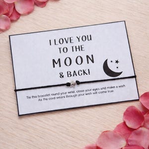 May include: A black and white card with the text "I LOVE YOU TO THE MOON & BACK!" and a crescent moon and star graphic. A black string bracelet with a star charm is attached to the card. Pink flower petals surround the card.