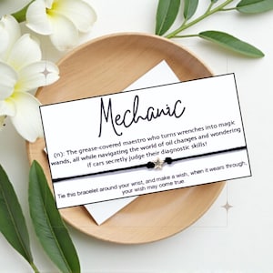 May include: A black string bracelet with two gold star charms. A white card with black text reads "Mechanic (n): The grease-covered maestro who turns wrenches into magic wands, all while navigating the world of oil changes and wondering if cars secretly judge their diagnostic skills! Tie this bracelet around your wrist, and make a wish, when it wears through, your wish may come true."