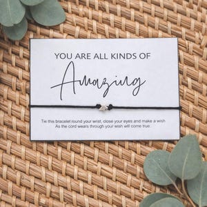 May include: A black cord bracelet with a silver star charm, displayed on a white card with the words "YOU ARE ALL KINDS OF Amazing". The card also includes instructions. The background is a woven basket.