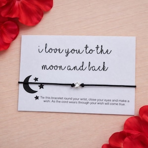 May include: A black string bracelet with a silver star charm, presented on a white card. The card features the phrase "i love you to the moon and back" and instructions. The design includes a crescent moon and stars.