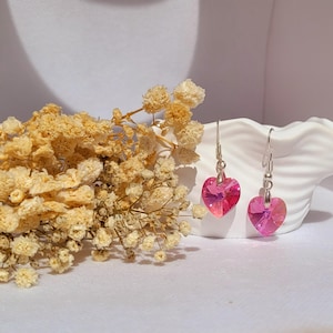 Puede incluir: Un par de pendientes en forma de corazón con un degradado de color rosa. Los pendientes cuelgan de ganchos plateados. Flores amarillas secas y un plato blanco en forma de concha en el fondo.