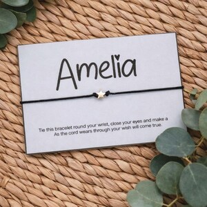 May include: A black cord bracelet with a small gold star charm, presented on a card with the name "Amelia" in black script. The card includes a message about making a wish. The background is a woven texture with green leaves.