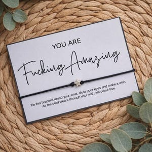May include: A black string bracelet with a silver star charm is displayed on a white card with the words "YOU ARE Fucking Amazing" in black script. The card also includes a message about making a wish. The background is a woven texture.