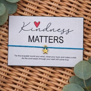 May include: A card with the words "Kindness Matters" and a blue string bracelet with a gold star charm. The card includes instructions to tie the bracelet and make a wish. The background is a woven wicker basket.