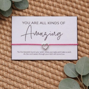 May include: A white card with the words "YOU ARE ALL KINDS OF Amazing" in black script. A pink string bracelet with a silver heart charm is attached. The card includes instructions.