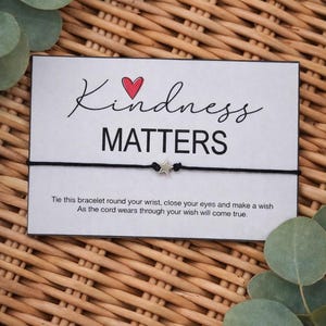 May include: A black cord bracelet with a silver star charm is displayed on a white card. The card reads "Kindness Matters" with a red heart and script font. The card is surrounded by eucalyptus leaves on a woven background.