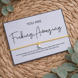 May include: A grey card with the words "YOU ARE Fucking Amazing" in black script. A yellow string bracelet with a silver star charm is attached. The text "Tie this bracelet..." is below the main text. The background is a woven texture with green leaves.