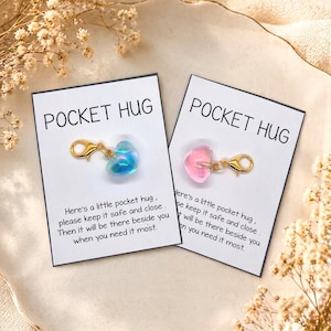 Peut inclure: Deux cartes avec le texte "POCKET HUG" et un charm en forme de cœur. Un charm est bleu, l'autre rose, tous deux avec des fermoirs dorés. Les cartes comprennent un message sur la sécurité et la proximité du câlin.