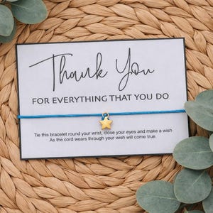 May include: A card with the words "Thank You" and "FOR EVERYTHING THAT YOU DO" in black script. A blue string bracelet with a gold star charm is attached to the card. The card is on a woven background with green leaves.