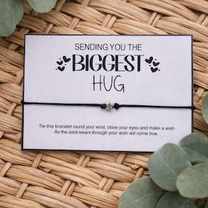 May include: A black cord bracelet with a silver star charm is presented on a white card. The card reads "SENDING YOU THE BIGGEST HUG" with a message below. The background is a woven wicker pattern with green leaves.