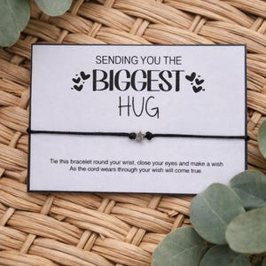 May include: A black cord bracelet with a small silver star charm is displayed on a white card. The card reads "SENDING YOU THE BIGGEST HUG" with a message below. The background is a woven wicker basket with green leaves.