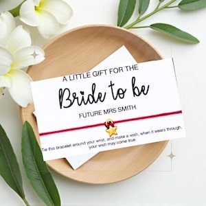 Op de afbeelding: Een rood koordarmband met een gouden sterbedel en een witte kaart met de tekst "Een klein cadeau voor de aanstaande bruid Toekomstige mevrouw Smith". De kaart zegt ook "Bind dit armbandje om je pols en wens iets, wanneer het versleten is, zal je wens uitkomen."
