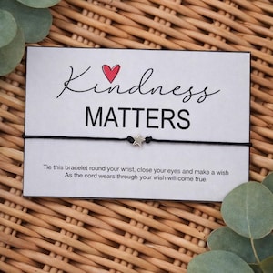 May include: A black cord bracelet with a silver star charm is displayed on a white card. The card reads "Kindness Matters" with a red heart and script font. The card is surrounded by eucalyptus leaves on a woven background.