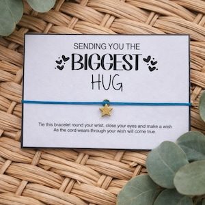 May include: A white card with the text "SENDING YOU THE BIGGEST HUG" and a blue string bracelet with a gold star charm. The card includes the text "Tie this bracelet round your wrist, close your eyes and make a wish. As the cord wears through your wish will come true."