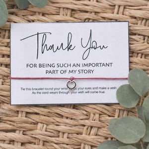May include: A white card with the words "Thank You" in elegant script, accompanied by the text "FOR BEING SUCH AN IMPORTANT PART OF MY STORY." A red string bracelet with a silver heart charm is attached. The card is on a woven background.
