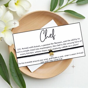 May include: A black string bracelet with a gold star charm, presented on a white card with the word "Chef" and a definition. The card is placed on a wooden plate, with white flowers and green leaves in the background.