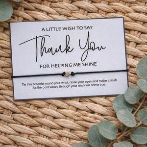 May include: A black cord bracelet with a silver star charm is displayed on a card that reads "Thank You". The card also says "For helping me shine" and includes instructions. The background is a woven, light brown surface with green leaves.