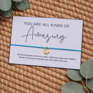 Op de afbeelding: Een kaart met de woorden "YOU ARE ALL KINDS OF Amazing" en een blauwe touw armband met een gouden ster bedel. De kaart bevat ook instructies. De achtergrond is een geweven textuur met groene bladeren.