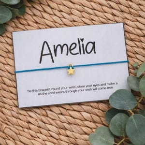 May include: A card with the name "Amelia" in black script, a gold star charm, and a teal cord bracelet. The card has text that reads, "Tie this bracelet round your wrist, close your eyes and make a wish. As the cord wears through your wish will come true."