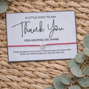 May include: A card with the words "Thank You" and a heart charm bracelet. The card says "A little wish to say Thank You for helping me shine." The bracelet is red with a silver heart charm. The background is woven.
