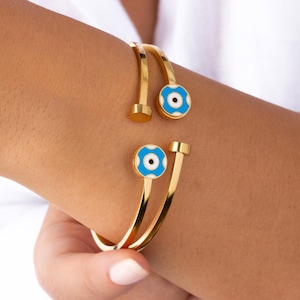May include: Two gold bangle bracelets with blue and white evil eye charms. The bracelets are open and have a hinged closure.