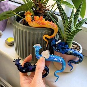 Könnte beinhalten: Eine Sammlung handgefertigter Drachenfiguren in verschiedenen Farben. Die Drachen sind in Blau-, Gelb- und Lilatönen gehalten und mit Wolken- und Sterndetails versehen. Sie werden in der Nähe von Topfpflanzen auf einer Fensterbank ausgestellt und zeigen ihr skurriles Design.