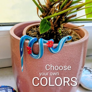 Peut inclure: Un tuteur de plante en forme de serpent bleu et blanc tourbillonnant avec des accents de fleurs rouges, enroulé autour d'un pot de fleurs rose. Le pot contient une plante verte. Le texte sur le pot indique "Choose your own COLORS".