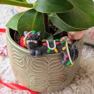 Op de afbeelding: Een zwart kattenbeeldje met een slinger van kleurrijke kerstverlichting, rustend in een decoratieve groene keramische pot met een plant. De pot is omgeven door kerstversieringen en een rood lint.
