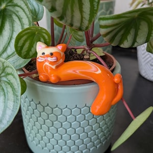May include: An orange cat figurine decorates a light green hexagonal pot holding a watermelon peperomia plant. The cat has white and orange markings. The plant has round green leaves with silver stripes.