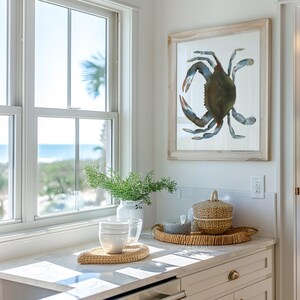 May include: A white kitchen counter with a window overlooking the ocean. There is a vase with green plants, three white bowls, and a woven tray with a woven basket and a glass container. A framed watercolor painting of a blue crab hangs on the wall.