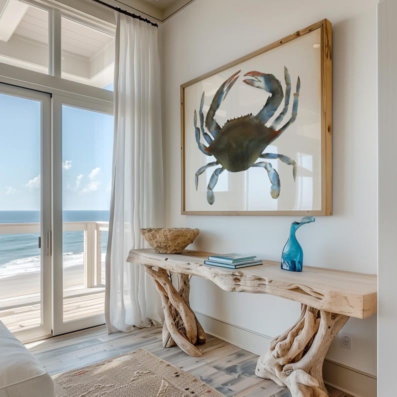 May include: A driftwood console table with a blue and brown crab painting above it. The table has a natural, rustic look and is decorated with a blue glass vase and a stack of books. The table is in front of a window with a view of the ocean.