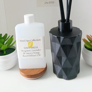 Puede incluir: Un frasco difusor de varillas negro con diseño geométrico y tres varillas negras, una botella de recambio de plástico blanco con la etiqueta Hotel Spa Collection HAPPINESS Bergamot, Lavender & Sweet Orange Reed Diffuser Oil y dos pequeñas suculentas verdes en macetas blancas.