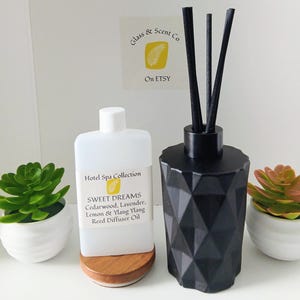 Könnte beinhalten: Eine weiße Kunststoffflasche mit Hotel Spa Collection Sweet Dreams Duftöl für Diffusoren mit Zedernholz, Lavendel, Zitrone und Ylang-Ylang, neben einem schwarzen geometrischen Diffusorgefäß mit schwarzen Stäbchen und zwei kleinen künstlichen Sukkulenten.