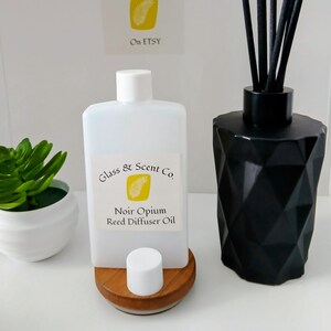 Könnte beinhalten: Eine weiße Kunststoffflasche mit dem Etikett Glass & Scent Co. Noir Opium Reed Diffuser Oil, ein schwarzer geometrischer Glasdiffusor mit schwarzen Stäbchen und eine kleine grüne Sukkulente in einem weißen Topf auf einem Holzsockel.