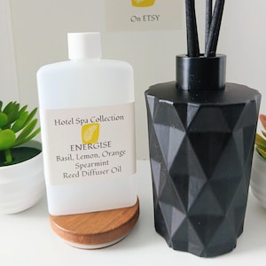 Könnte beinhalten: Eine weiße Kunststoffflasche mit Hotel Spa Collection Energise Duftöl für Diffuser mit einem Etikett für Basilikum, Zitrone, Orange und grüne Minze, neben einem schwarzen, geometrischen Glasdiffuser mit schwarzen Stäbchen und Holzsockel.