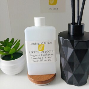 Könnte beinhalten: Eine weiße Kunststoffflasche mit Hotel Spa Collection Refresh & Focus Duftöl für Diffuser mit Bergamotte, Eukalyptus, Lavendel und Zitrone. Sie steht auf einem Holzsockel neben einem schwarzen, geometrischen Diffuser-Gefäß und einer kleinen Sukkulente.
