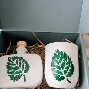 May include: A white ceramic bottle with a cork stopper and a matching white ceramic cup, both featuring a hand-painted green leaf design. The items are nestled in a gift box filled with brown paper shred, showcasing a clean and simple aesthetic.