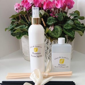 Puede incluir: Un frasco de spray ambiental blanco y un frasco transparente de aceite para difusor de varillas, ambos con la etiqueta Glass & Scent Co. Peonies. El set incluye un manojo de varillas de madera clara y otro de varillas negras, atados con una cinta color crema.