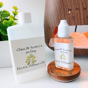 May include: A large translucent plastic bottle and a smaller clear glass bottle, both with white caps and labels reading Glass & Scent Co. on Etsy Electric Diffuser Oil with a green leaf graphic. The bottles are positioned near a wood-grain diffuser.