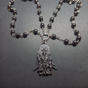 May include: A black beaded necklace with a silver pendant featuring a detailed depiction of a winged figure. The pendant is attached to a silver chain. The necklace has two silver circular charms with a star design.