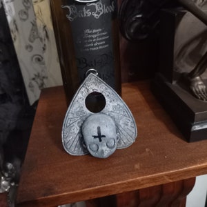 Peut inclure: Un pendentif en argent en forme de chauve-souris avec les mots "The Spirit of Wicked" inscrits dessus. Le pendentif est suspendu à une chaîne et est attaché à une bouteille de vin étiquetée "Bats Blood". Un pendentif de planche Ouija en argent avec un design de crâne et d'os croisés est également représenté.