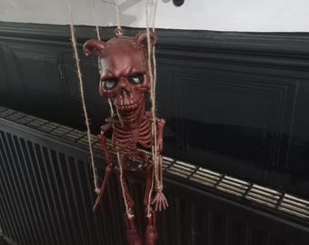 Może przedstawiać: Marionetka szkielet diabła w kolorze bordowym z rogami, zawieszona na sznurkach. Lalka ma szczegółową czaszkę i klatkę piersiową, z małymi dłońmi i stopami. Wisi na czarnym ozdobnym wsporniku, sugerując motyw Halloween lub gotycki.