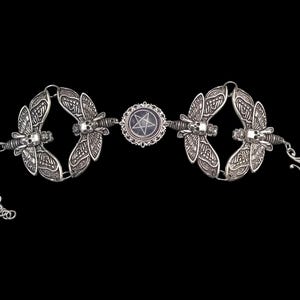 May include: Silver-toned bracelet featuring ornate dragonfly designs with skull accents. The bracelet is connected by a central circular element with a pentagram. The bracelet has a chain and hook closure, set against a black background.