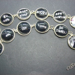 May include: A black and silver bracelet with round charms featuring white text. The charms have phrases like "You Can't", "Loving You", "Cause Your Roots Are Showing", "Like Loving The Dead".