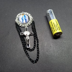 Op de afbeelding: Een religieuze broche met een zilverkleurig kruis met een Jezusfiguur en een cirkelvormige hanger met een blauw-witte afbeelding. Zwarte ketting hangt van de broche. Een gele AA-batterij ligt naast de broche.