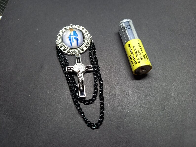 Op de afbeelding: Een religieuze broche met een zilverkleurig kruis met een Jezusfiguur en een cirkelvormige hanger met een blauw-witte afbeelding. Zwarte ketting hangt van de broche. Een gele AA-batterij ligt naast de broche.