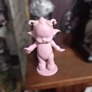 May include: A pink figurine of a small, horned creature with a human-like body and a whimsical expression. The figurine is standing on a round base.