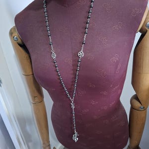 K&ouml;nnte beinhalten: Eine lange Halskette mit dunklen Metallperlen auf einer silberfarbenen Kette. Das Design umfasst zwei kreisf&ouml;rmige, silberfarbene Pentagramm-Anh&auml;nger, die in die Perlenarbeit integriert sind, sowie einen Anh&auml;nger am unteren Ende der Kette.