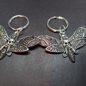 Może przedstawiać: Dwa srebrne breloki z detalicznym wzorem ćmy z czaszką. Ćmy mają skomplikowane wzory skrzydeł i fakturowane ciało.